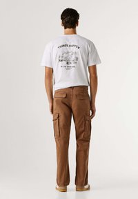 Wit t-shirt met een grafische print op de achterkant, gecombineerd met bruine cargobroeken met meerdere zakken en een losse pasvorm. Casual schoeisel zichtbaar.