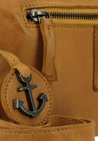 Borsa in pelle color cuoio con etichetta rotonda che presenta un emblema di ancora, accessori in argento e cuciture dettagliate lungo i bordi. Zip visibile.