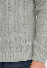 Gray knitted sweater featuring cable patterns, ribbed cuffs, and hem. Includes a small label at the bottom right with brand details.