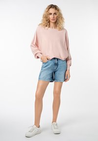 Hellrosa übergroßer Pullover mit überschnittenen Schultern, kombiniert mit blauen Jeansshorts. Das Modell trägt weiße Sneakers. Einfaches, lässiges Outfit.