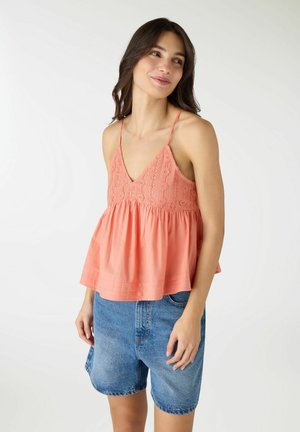 Donna che indossa un top senza maniche rosa corallo con dettagli in pizzo e pantaloncini di jeans larghi, in piedi davanti a uno sfondo bianco semplice.