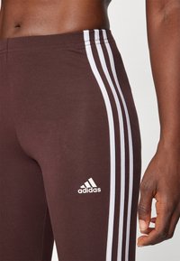 Hnědé sportovní legíny s bílými trojicí pruhů po stranách a logem Adidas na dolní části levé nohy. Hladká textura.