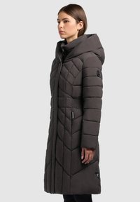 Mujer vestida con un largo abrigo de invierno acolchado de color gris oscuro con una gran capucha, de pie de perfil contra un fondo claro y liso.