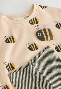 Bež otroško oblačilo z odtiskanimi čebelami in 3D čebeljo aplikacijo, združeno s črno-belimi črtastimi hlačami.