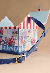 Curea albastră a genții cu cataramă aurie în fața unei cutii mici deschise, concepută ca un magazin de flori cu verandă în dungi roșii și albe și ilustrații florale.