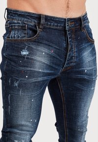 Úzké džínové kalhoty tmavě modré barvy s prošoupanými detaily a skvrnami od barvy v bílé a červené. Klasický design s pěti kapsami a oranžovým prošitím.