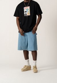 Czarna koszulka z kolorową grafiką, jasnoniebieskie szorty dżinsowe, beżowe buty i białe skarpetki z czarnym napisem. Casualowy, luźny krój.