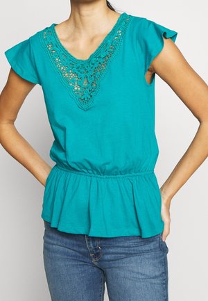 Kobieta ubrana w morski zielony top bez rękawów z koronkowymi zdobieniami przy dekolcie, zebrany w talii, w połączeniu z niebieskimi dżinsami.