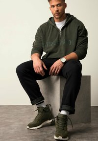 Felpa con zip verde e testo "STREETPOET", jeans neri e sneaker alte verdi. Modello seduto su un piedistallo in cemento, sfondo neutro.