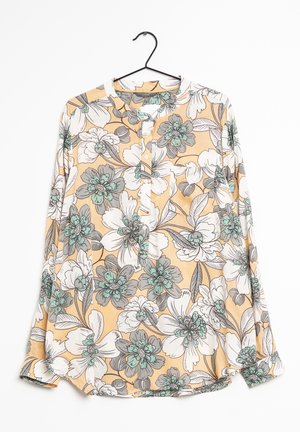 Dlouhý rukáv květinová halenka s broskvovým pozadím, bílými a tyrkysovými květy, s knoflíkovou légou vpředu, zavěšená na černém ramínku na bílém pozadí.