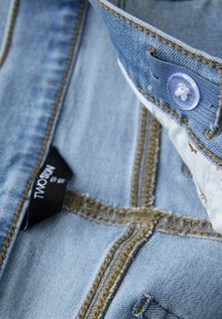 Un tissu en denim bleu clair présente un col classique, un bouton clair, des coutures marron contrastantes et une étiquette noire portant la mention "TWO."