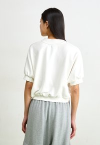 Sweatshirt branco de manga curta com bainha e punhos franzidos, feito de material macio. Usado sobre calças cinzentas largas.