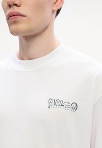 Bílý bavlněný tričko s texturovaným, lesklým černým logem na levé straně hrudi. Límec je kulatý a látka vypadá měkce.