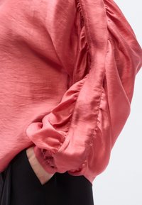 Κοραλλένιο πουκάμισο με μακριά μανίκια και φουσκωτά μανίκια, με απαλή υφή και σούρες στα μανίκια, φορεμένο πάνω από μαύρο παντελόνι.