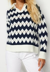 Donna che indossa un maglione lavorato a maglia blu navy e bianco con un motivo a rombi, orlato in bianco, e pantaloni bianchi, in posa davanti a uno sfondo semplice.