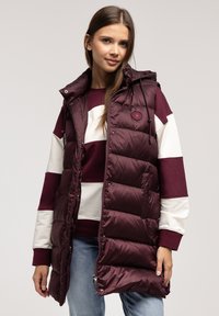 Bordowy kamizelka puchowa z kapturem, z błyszczącym wykończeniem, bocznymi kieszeniami i zapięciem na zamek. Noszona na paskowany sweter i niebieskie dżinsy.