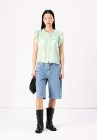Blusa verde claro, com babados e fecho de botão, combinada com calções de ganga azuis. A modelo usa botas altas pretas até ao joelho e segura uma mala preta.
