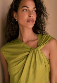 Groene, mouwloze jurk met een geknoopte voorkant, zachte textuur en gedrapeerd stof, waardoor een schouder zichtbaar is. Krullend haar en neutrale make-up.