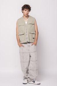 Light khaki sleeveless vest with four front pockets, paired with loose gray pants featuring a stitched pattern. Worn with white and gray sneakers.
