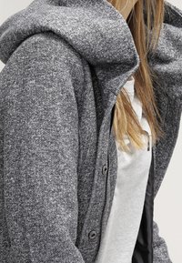 Felpa grigia con trama, con ampio colletto piegato e cerniera frontale. Presenta dettagli con bottoni e un effetto a strati sopra una maglietta chiara.