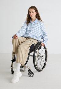 Randig blå och vit långärmad polo-tröja, beige baggy byxor, vita sneakers, sittande i en rullstol med svarta hjul.