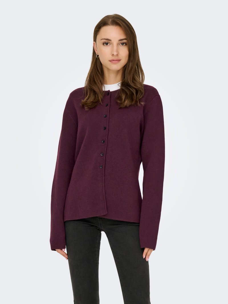 Ung kvinde, der vender fronten mod, iført en knappet blommefarvet cardigan over en hvid top og sorte jeans mod en ensfarvet lys baggrund.