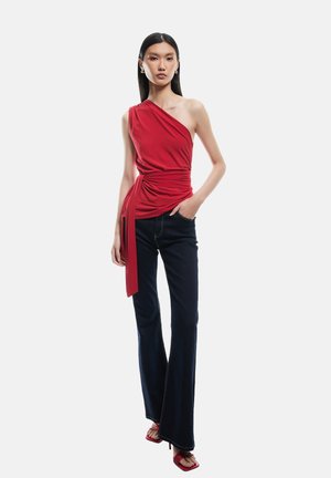 Top rojo de un solo hombro con tejido fruncido y detalle de lazo, combinado con jeans oscuros de campana. La modelo lleva sandalias rojas con tiras.
