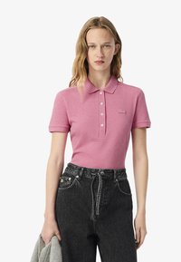 Polo rosa con colletto, tre bottoni e logo ricamato. Abbinato a jeans in denim scuro a vita alta e un maglione grigio sul braccio.