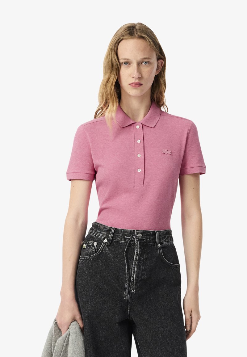 Polo rosa con colletto, tre bottoni e logo ricamato. Abbinato a jeans in denim scuro a vita alta e un maglione grigio sul braccio.