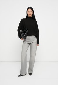 Schwarzer Rollkragenpullover, graue weite Jeans und schwarze High-Heels. Model hält eine schwarze texturierte Clutch. Einfacher Hintergrund.