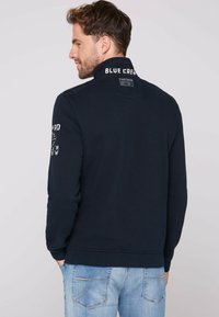 Felpa navy con collo alto, polsini e vita a coste. Presenta il testo "BLUE CREW" e una toppa sul retro, con una texture liscia.