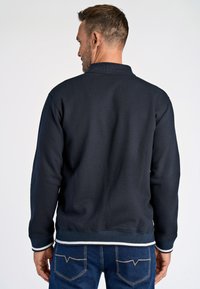 Felpa blu navy con colletto alto, polsini e orlo a coste. Presenta strisce bianche a contrasto sull'orlo. Abbinata a jeans in denim blu.