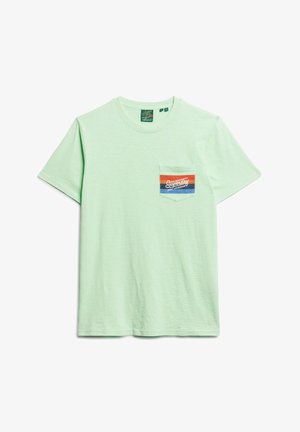 Camiseta de algodón de manga corta en verde claro. Presenta un bolsillo en el pecho izquierdo con una etiqueta gráfica multicolor. Textura suave.