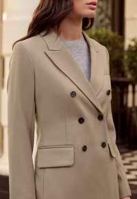 Sacou bej cu două rânduri de nasturi negri, revere cu decupaj și buzunar la piept. Material texturat și croială ajustată.