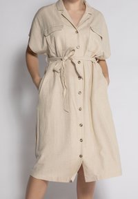 Vestito in lino beige con cintura, colletto, maniche corte, due tasche sul petto e chiusura con bottoni frontali. Texture liscia.
