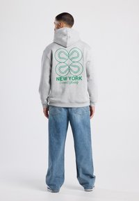 Felpa grigia con una grafica verde che presenta le parole "NEW YORK Tennis Society" e un design circolare sul retro. Indossata con jeans a gamba larga.