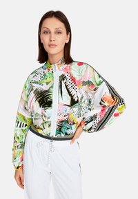 Colorful bomber jacket with tropical floral prints, featuring a high collar, zip closure, and elastic cuffs. Paired with white trousers.