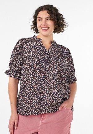 Smilende kvinde med kort krøllet hår iført en navybluse med lyserøde blomster og lyserøde bukser, stående med den ene hånd i lommen.