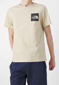 Camiseta de algodón beige con un logo cuadrado negro en el pecho con texto blanco y un diseño curvado icónico. Mangas cortas y escote redondo.
