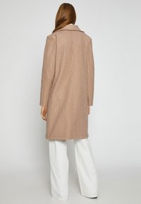 Beige Wollmischungsjacke mit Reverskragen, langen Ärmeln und geradem Schnitt, kombiniert mit weißen weiten Hosen, von hinten betrachtet.