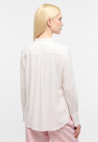 Weiße Bluse mit langen Ärmeln, die eine weiche, glatte Textur, einen Kragen und einen Rückenfalte aufweist. Kombiniert mit hellrosa Hosen.