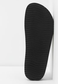 Semelle de chaussure noire avec des rainures horizontales appuyée contre un mur et un sol blancs.