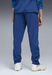Blaue Sporthosen aus glattem Stoff mit tailliertem Design, elastischen Bündchen und seitlichen roten Akzenten, getragen mit weißen Fußballschuhen.