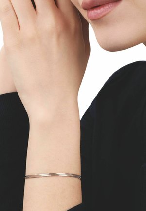 Poignet féminin avec un bracelet fin en or rose, main reposant près du menton, lèvres visibles, sur fond blanc et vêtement noir.