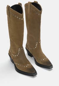 Smeđe suede cowboy čizme s špicastim prstima, dekorativnim metalnim zakovicama duž šavova i masivnim crnim potplatom.