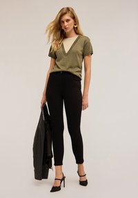 Top verde oliva de manga corta con escote en V y acentos beige, combinado con jeans ajustados negros y zapatos de tacón altos negros, sosteniendo una chaqueta de cuero.