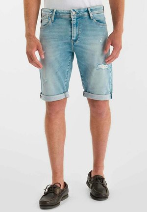 Uomo che indossa pantaloncini di jeans azzurri chiari risvoltati con dettagli consumati e scarpe stringate marrone scuro, in piedi su uno sfondo bianco.