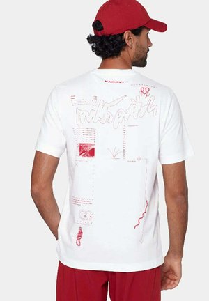 Hombre con camiseta blanca con diseños abstractos rojos y gorra roja de espaldas, de pie frente a un fondo liso.