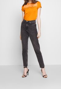 Oranžové tričko s krajkovým detailem na přední straně, spárované s černými riflemi s vysokým pasem a černými páskovými sandály na vysokém podpatku.