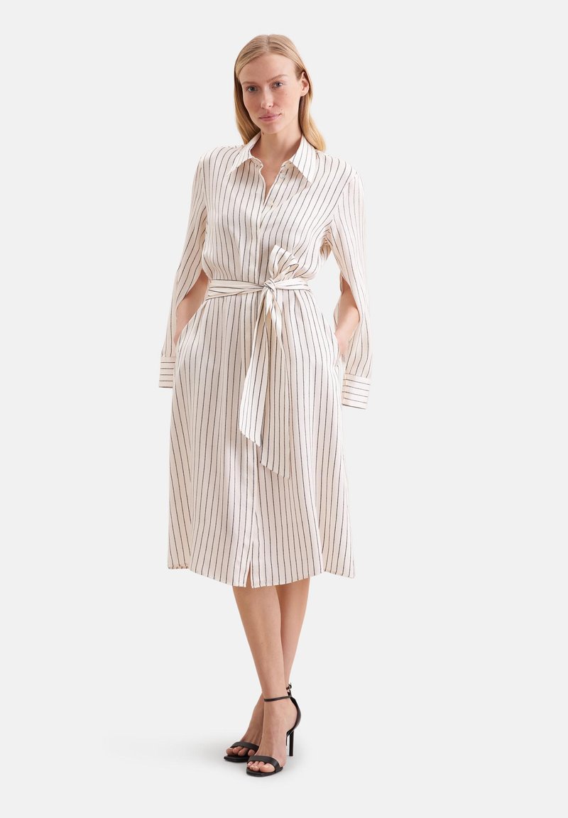 Femme portant une robe midi blanche à rayures verticales noires, une ceinture nouée à la taille, des manches fendues et des sandales noires à talons hauts, debout sur un fond blanc.